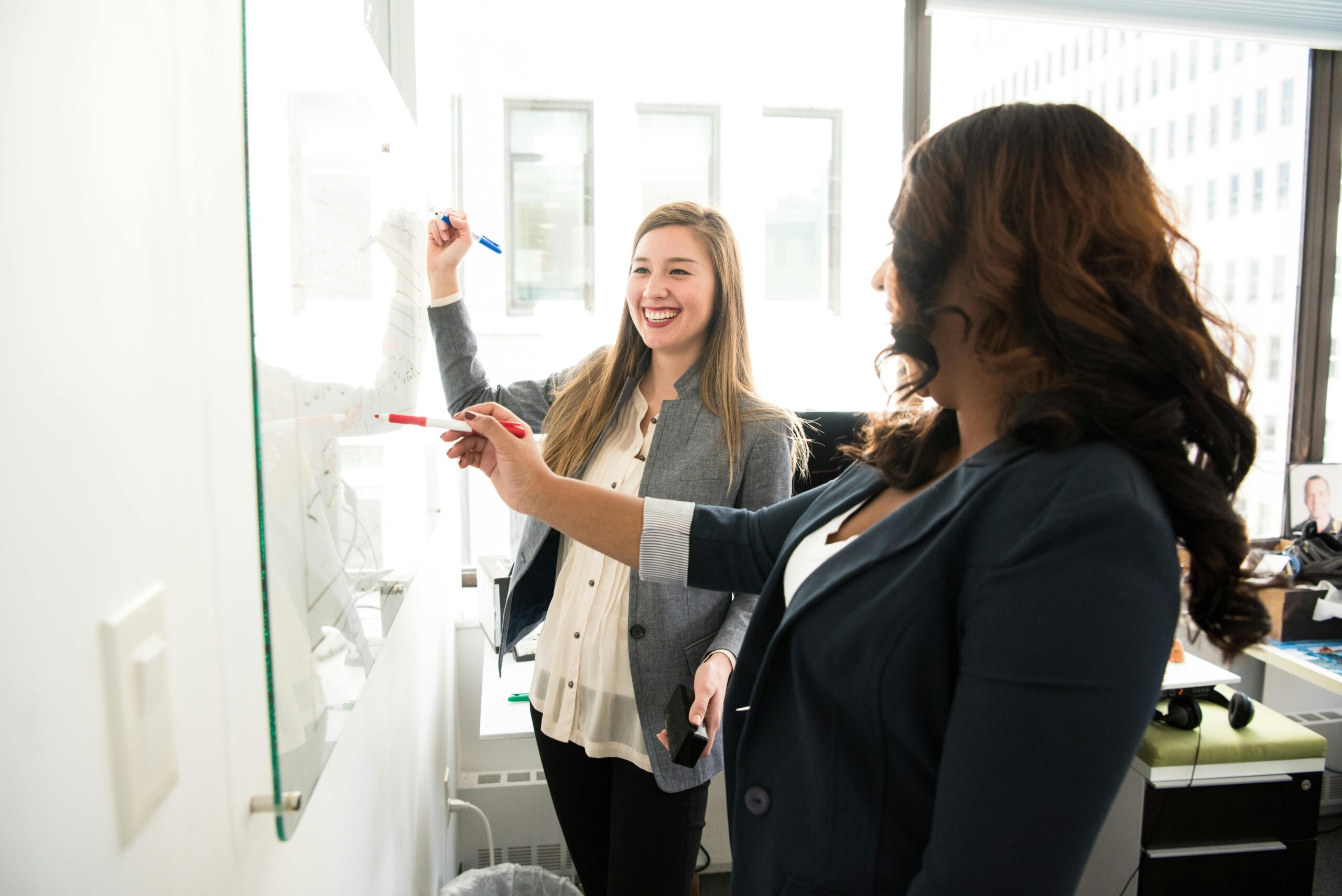 Two professional women discussing ideas on a whiteboard in a modern office setting